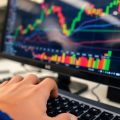 Mână pe tastatură, monitor cu grafice financiare.